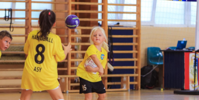 Grafika przedstawia dziewczynę na hali sportowej rzucającą piłką